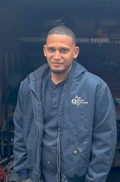 The image shows a smiling serviceman wearing an "Air Quality Systems" jacket, posing in front of equipment.