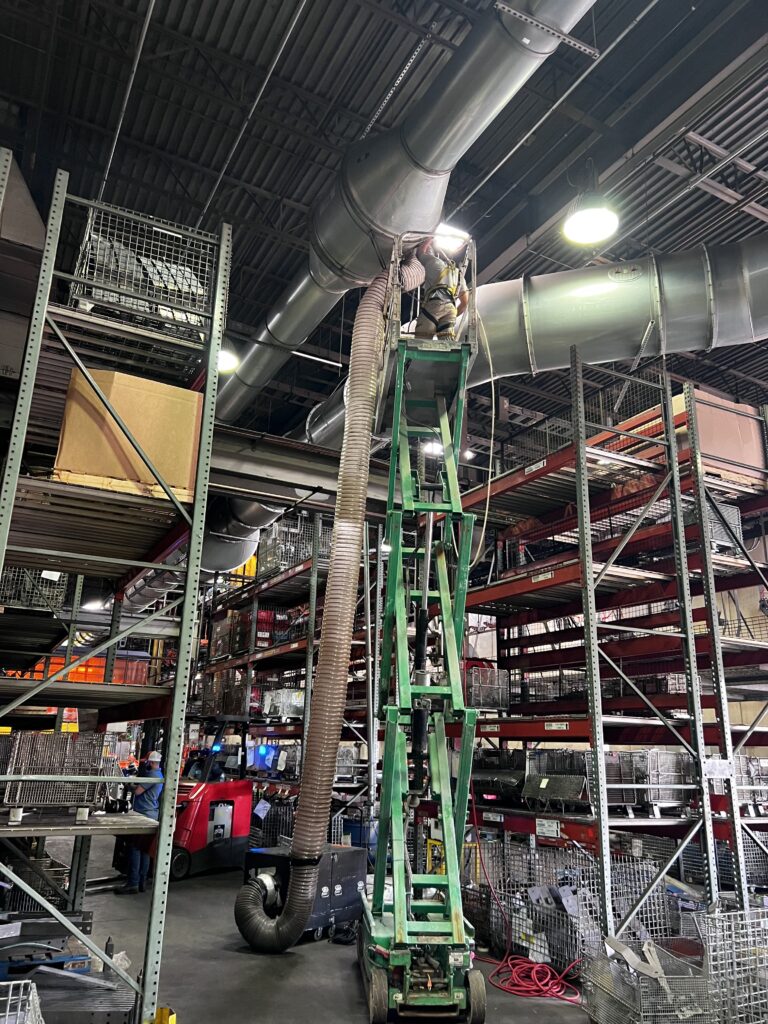 Worker on tall ladder cleaning air ducts in large industrial warehouse with racks of supplies