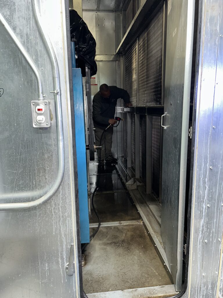 Man cleaning narrow air duct tunnel with vacuum hose and brush attachment in enclosed metal space.