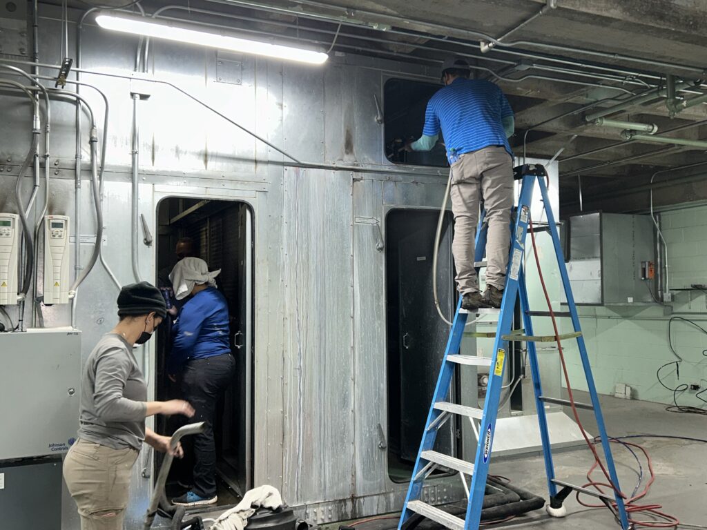 Commercial air duct cleaning technicians using ladders to access and clean an HVAC system in a building.