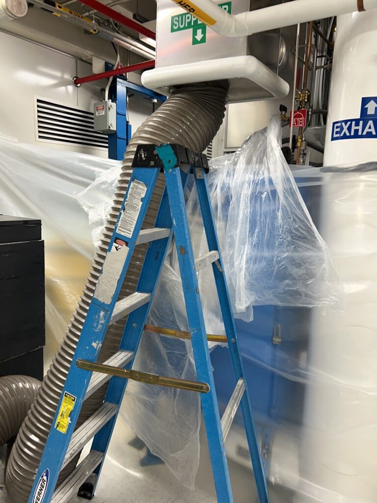 Commercial air duct cleaning equipment, including a blue ladder and plastic sheeting, in a mechanical room