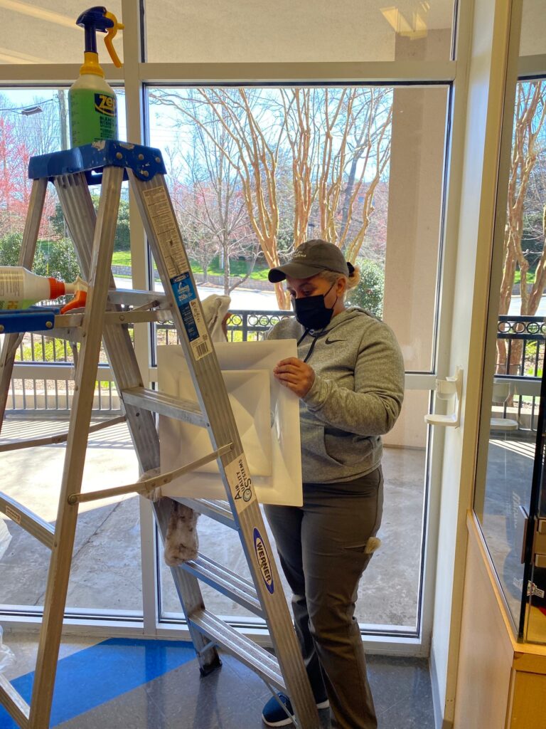 Commercial air duct cleaning technician applying sanitizer spray to residential ductwork on ladder